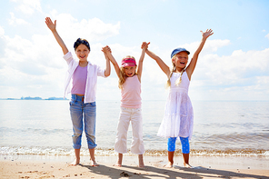 Carefree girls holding by hands and raising them while enjoying sunny day on the beach