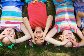 Directly above view of little girls lying on green grass in park and making funny faces with their fingers around eyes