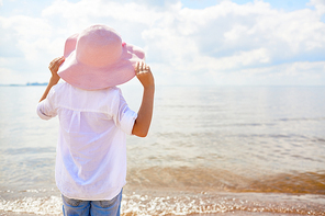 Back view of little girl in pink hat standing on beach in front of water