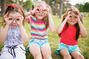 Cute little girls making funny faces with fingers around eyes while sitting on green grass in park