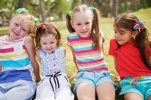 Close-up view of cheerful little girls in summer clothes sitting on green grass in park and 