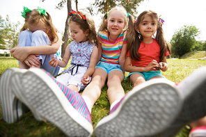 Low angle view of pretty little girls in sneakers resting on green lawn in park on a warm summer day