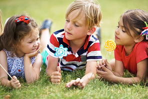 Close-up view of cute kids lying on green grass in park, holding lollipops and chatting on wonderful summer day