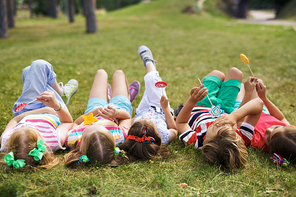 Group of children in colorful summer wear lying on their backs on green lawn in park and eating lollipops