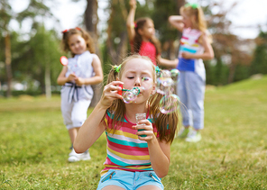 Pretty little girl with ponytails sitting on grass and blowing soap bubbles on background of her friends in park