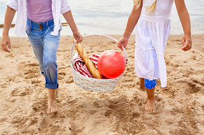 Little friendly girls carrying basket with tablecloth, red ball and crusty baguette while walking down sandy beach