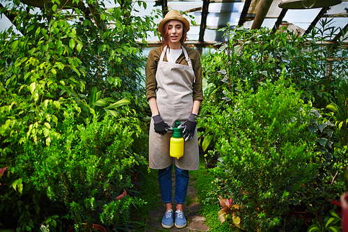Portrait of beautiful young woman wearing straw hat and apron enjoying work in glasshouse, standing full height and  smiling between tree shrubs