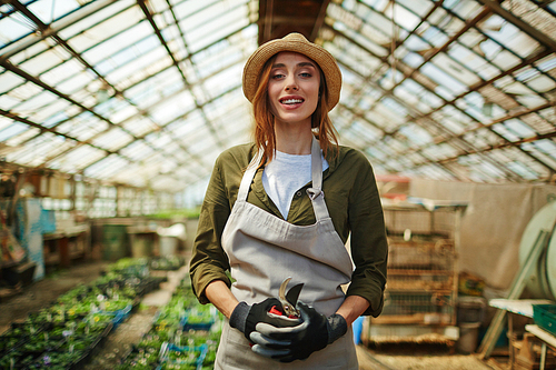Portrait of smiling beautiful woman wearing straw hat enjoying working with plants in glasshouse, standing  in flower nursery