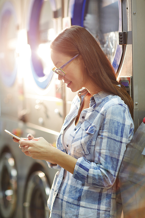 Happy young woman texting in cellphone by washing-machine