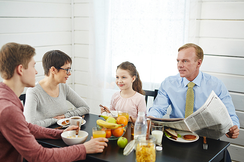 Peaceful breakfast in bosom of family: red-haired father with newspaper in hands talking to his teenage son, little daughter and her short-haired mother enjoying delicious food