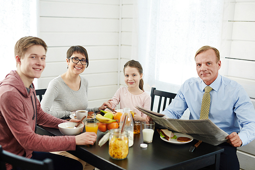 Loving family posing for photography with wide smiles while sitting at breakfast table full of delicious and healthy food, waist-up portrait