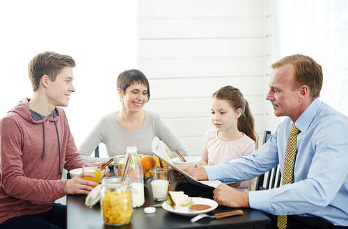 Family of four sitting around kitchen table and enjoying delicious breakfast, concentrated father reading newspaper together with his curious little daughter