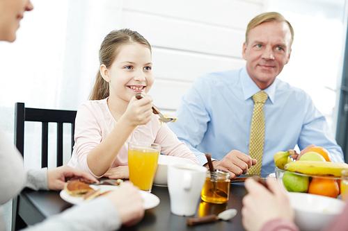 Waist-up portrait of pretty little girl holding spoon with cornflakes in hand and looking at her brother with smile, other family members also enjoying breakfast