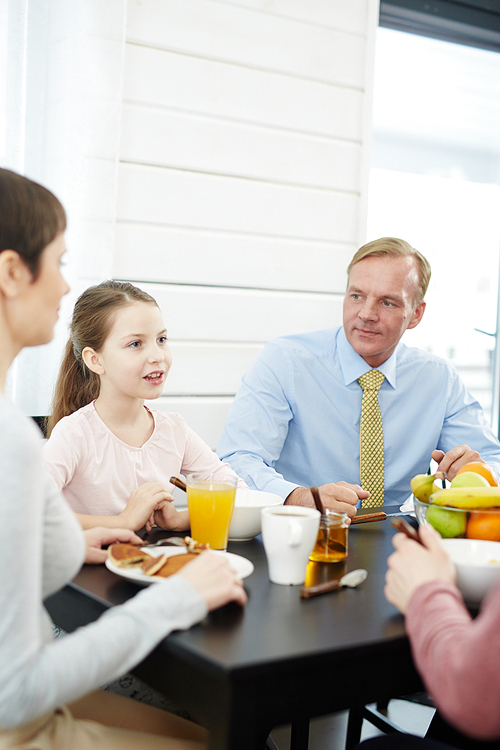 Portrait of fair-haired middle-aged man in shirt and tie refreshing himself with delicious breakfast and listening to his little daughter with curiosity