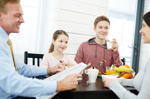 Happy middle-aged parents and their children starting new day with delicious breakfast, handsome father reading newspaper, waist-up portrait