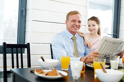 Businessman with newspaper and his daughter sitting by table in the kitchen