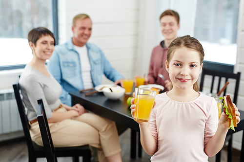 Waist-up portrait of pretty little girl  with smile while holding glass of orange juice in one hand and healthy sandwich in other, her family sitting at kitchen table