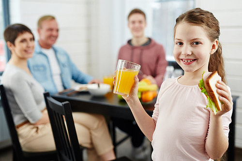 Portrait of primary age girl posing for photography with glass of orange juice and healthy sandwich, her family looking at her while sitting at dining table