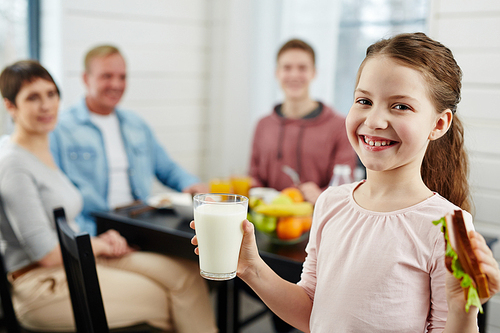 Cute little girl with ponytail holding glass of milk and tasty sandwich in hands while  with toothy smile, her family sitting at kitchen table