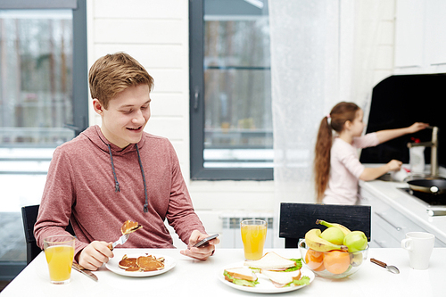Portrait of smiling teenage boy texting with his friend on smartphone while sitting in kitchen and eating pancakes, his little sister standing at sink and washing dishes