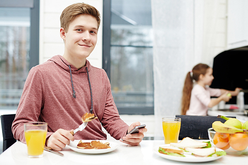 Waist-up portrait of handsome teenage boy  with wide smile while eating tasty pancakes at breakfast, his little sister standing at sink and washing dishes