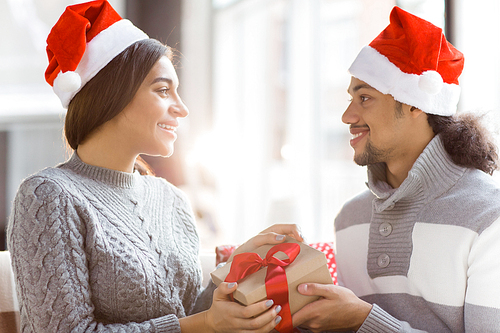 Happy young dates in Santa caps looking at one another while holding giftbox