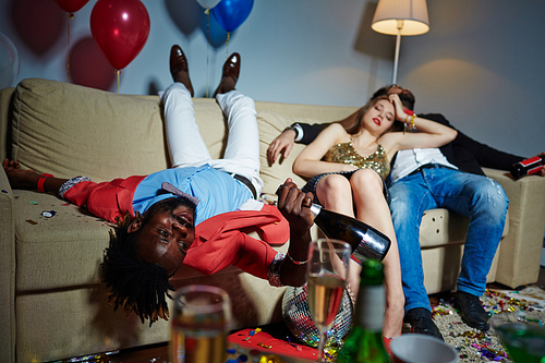 Group of glamorous friends relaxing on sofa after wild house party, they holding champagne and beer bottles in hands while having nap