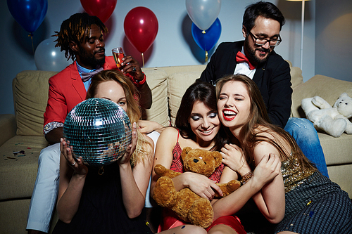 Well-dressed colleagues having New Year party: two men with bow ties sitting on comfortable sofa while three young women posing for photography