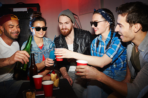 Group of smiling party people, men and women, drinking beer and raising glasses hanging out in night club