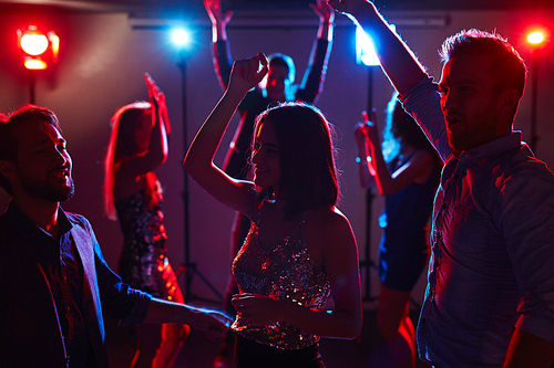 Group of dressed-up male and female students celebrating graduation in night club, dancing with hands in the air and enjoying party