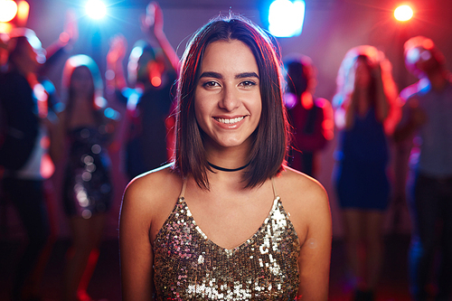 Portrait of pretty brown-eyed girl in shining top standing on dancing crowd background and smiling at camera cheerfully