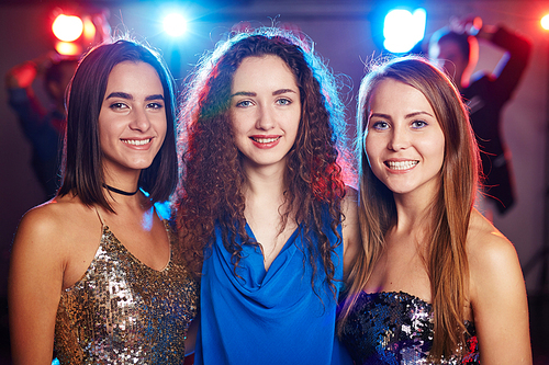 Portrait of three beautiful female students in fancy clothes standing together and smiling at camera happily while having graduation party in club