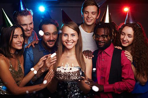 Group photo of dressed-up young people celebrating birthday of their female friend standing in the middle with cake and smiling happily