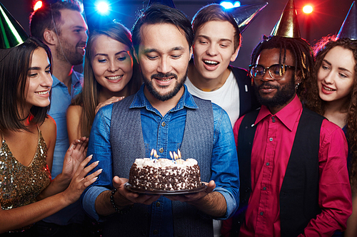 Young bearded man standing among close friends, holding birthday cake with candles and making a wish