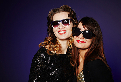 Waist-up portrait of two stylish women in sunglasses  with wide smiles while standing against dark background, studio shot