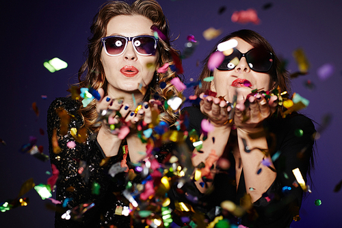 Having fun with best friend: two stylish women standing against dark background and blowing colorful confetti, waist-up group portrait