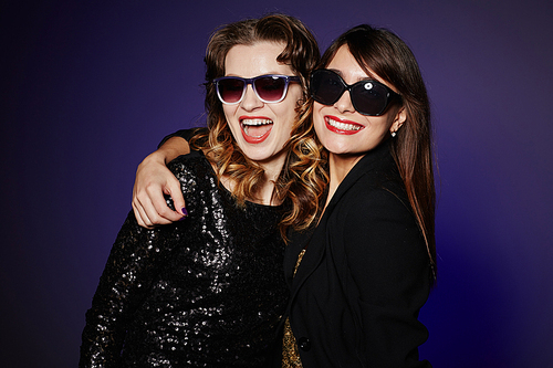 Waist-up portrait of joyful party goers wearing sunglasses posing for photography while standing against dark background