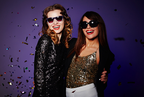 Two cheerful it girls gathered together in modern night club and  with charming smiles, colorful confetti falling on background