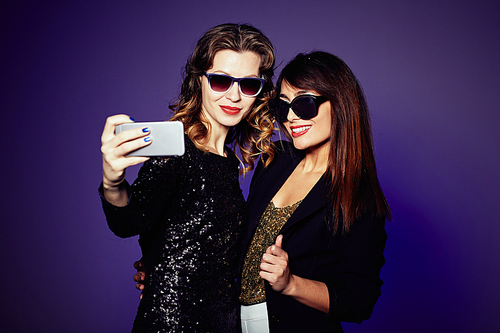 Stylish young women in sunglasses taking selfie on smartphone while standing against dark background