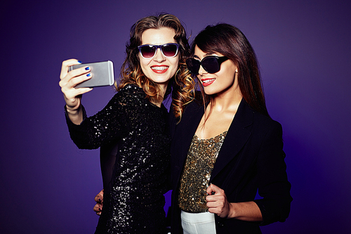 Attractive it girls in trendy sunglasses taking selfie on smartphone before starting to celebrate New Year, waist-up portrait