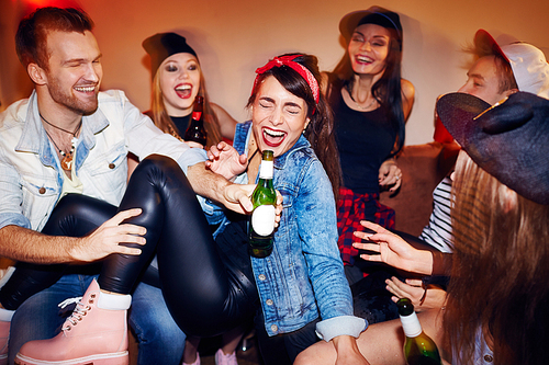 Group of trendy young people getting drunk at late night swag party, going crazy and laughing while sitting on sofa with beer bottles