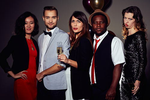 Group portrait of young friends wearing stylish clothes celebrating Christmas in night club, attractive young woman with dissatisfied facial expression holding champagne flute in hand