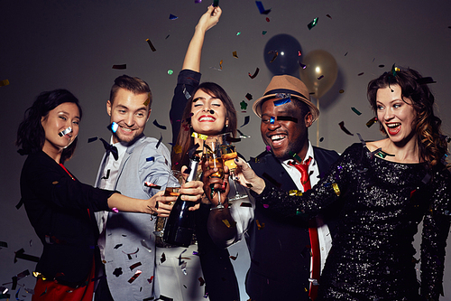 Having New Year party in nightclub: multi-ethnic group of friends dancing and posing for photography with champagne flutes in hands, colorful confetti falling on background