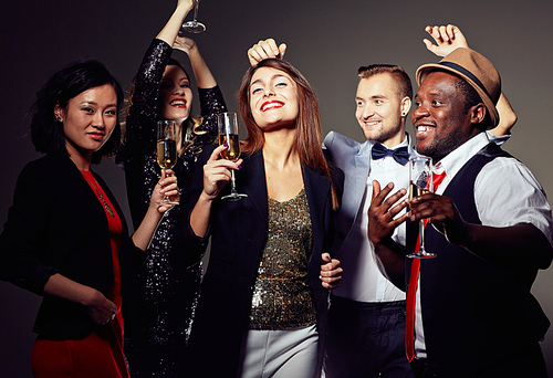 Multi-ethnic group of smiling friends in evening wear posing for photography while dancing with champagne flutes in hands