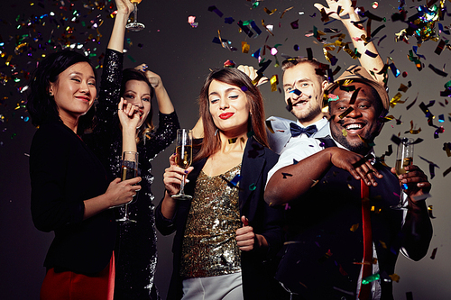 Joyful party at full speed: five smiling friend in evening wear dancing and drinking champagne at night club, colorful confetti falling, dark background