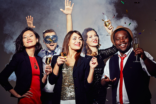 Enthusiastic stylish friends hanging out in trendy night club, they holding champagne flutes in hands, young bearded man in carnival mask dancing with hands up