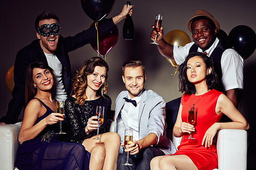 Group portrait of trendy young people sitting on sofa with champagne flutes in hands and posing for photography with wide smiles, two handsome men standing behind them