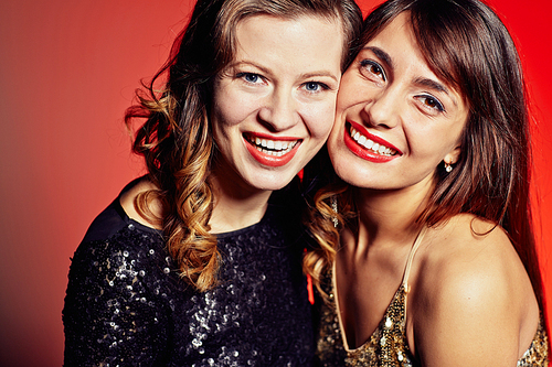 Head and shoulders portrait of pretty young women standing cheek to cheek against red background and  with toothy smiles
