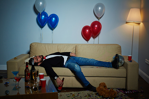 Completion of wild party: middle-aged man with beer bottle in hand sleeping on sofa, plastic cups, empty alcohol bottles and colorful confetti observed everywhere
