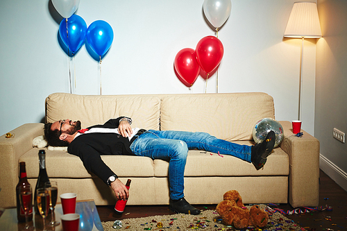 Smiling middle-aged man lying on couch and drinking beer from bottle in messy living room, empty alcohol bottles and plastic cups standing everywhere
